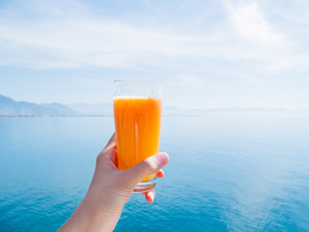 手与新鲜美味的新鲜榨果汁的成熟橙子在玻璃。平静的大海，地平线上有山脉。克姆,土耳其。图片下载