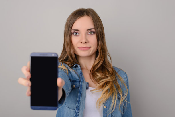 微笑的女子，棕色长发和牛仔裤衬衫显示空的黑色智能手机屏幕;在灰色背景上孤立图片下载