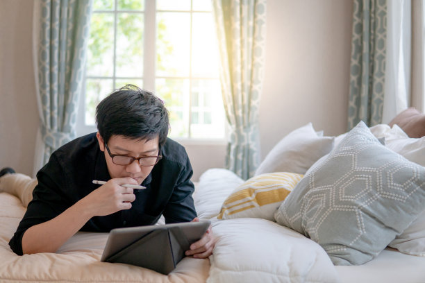 年轻的亚洲商人躺在舒适的卧室的床上使用数字平板电脑和笔。具有现代电子小玩意概念的家居生活方式图片下载