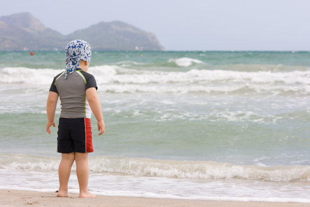 2岁的小男孩穿着泳衣在沙滩上。准备好游泳。看着海浪。图片下载