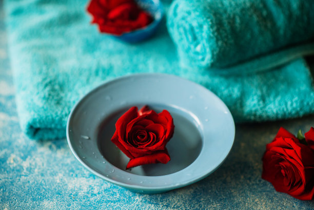 水疗和自我护理的概念:玫瑰水和红玫瑰花，旁边的毛巾在蓝色的背景。图片下载