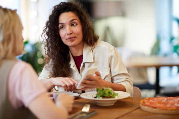 积极的年轻混血女士卷发坐在桌子上，向朋友展示智能手机上的照片，而他们在咖啡馆讨论生活事件图片下载