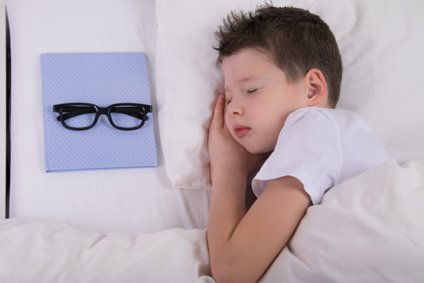这个男孩睡在一张白色的床上，旁边放着眼镜，以改善他的视力图片下载