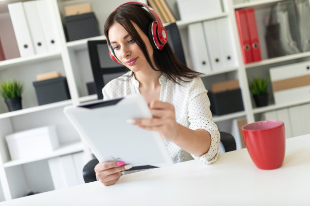 一个戴着耳机的小女孩坐在办公室的电脑桌前，手里拿着平板电脑。图片下载