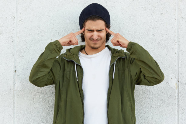 水平射击紧张的十几岁的男孩关闭是的，感到压力，保持两个食指在太阳鬓，戴着黑色的帽子和绿色的夹克，孤立在白色的背景，回忆信息图片下载