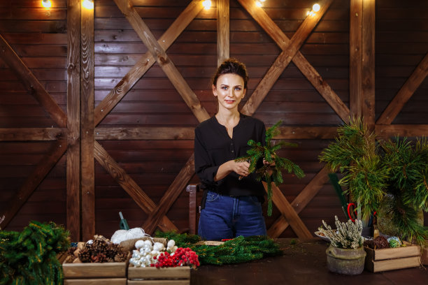 工作花店的女人与圣诞花环。年轻可爱的微笑女人设计师准备圣诞常青树花环图片下载