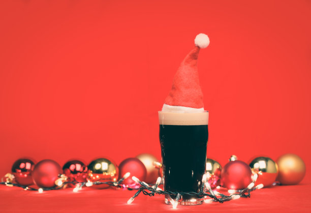 黑啤酒品脱玻璃杯与红色圣诞帽，圣诞装饰物和灯在红色背景色调图片下载
