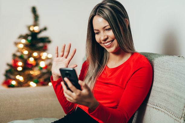 一名年轻女子坐在圣诞树旁的沙发上玩手机图片下载