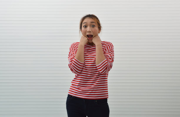 快乐和惊讶的年轻亚洲女人穿着红白条纹衬衫和牛仔裤手在她的脸颊在白色墙壁的背景，愉快的面部表情图片下载