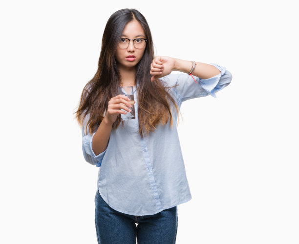 年轻的亚洲女人喝一杯水在孤立的背景与愤怒的脸，消极的迹象显示不喜欢拇指向下，拒绝的概念图片下载