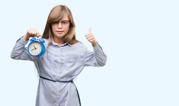 年轻的金发小孩抱着闹钟开心地笑着做ok的手势，用手指竖起大拇指，优秀的手势图片下载