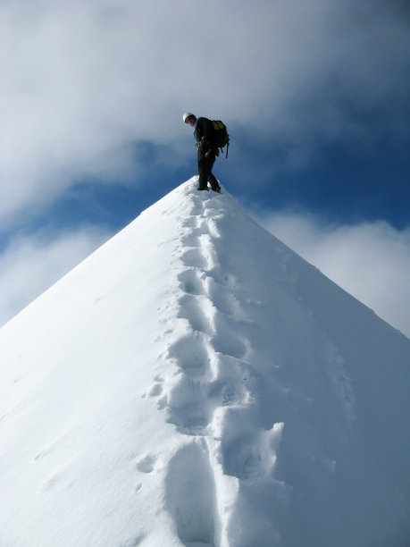 一个登山者，站在一个狭窄的、裸露的、有轨道向上的雪山山顶上图片下载
