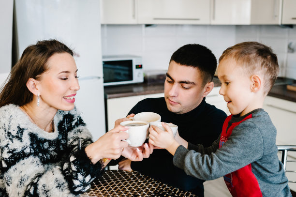 年轻的妈妈，爸爸和男孩在早上喝咖啡和茶。快乐的妈妈，爸爸和儿子在家里的厨房里吃早餐。家庭、饮食、人的观念。图片下载