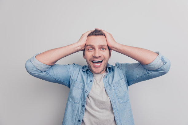 好看的照片好看的穿着的男人与张大的嘴瞪着眼睛握着双手在头上看相机站孤立在浅灰色的背景图片下载