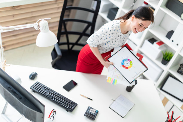 一个年轻的女孩站在办公室的电脑桌旁边，手里拿着文件夹和图表。图片下载