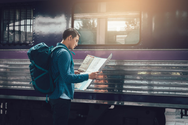 后视图的背包亚洲人拿着地图纸和看路线，而在火车站等待火车。图片下载