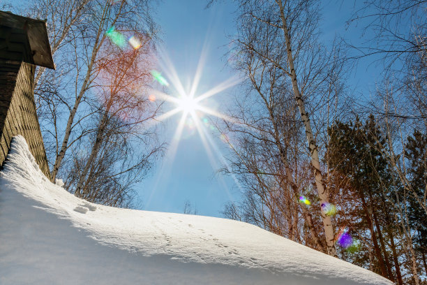 带有烟囱的乡村小屋屋顶上覆盖着一层厚厚的雪。蓝色的天空和太阳耀斑的背景。冬天在森林里的乡村小屋图片下载