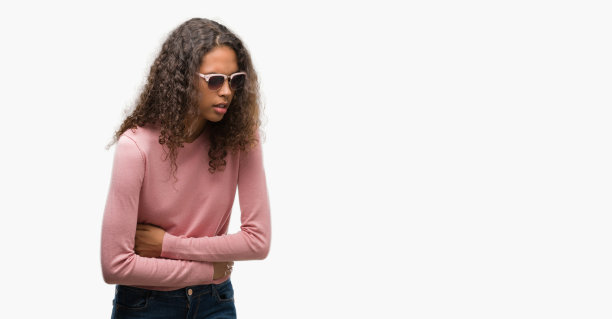 年轻漂亮的西班牙女子戴着墨镜，手放在肚子上，因为恶心，痛苦的疾病感觉不舒服。疼痛的概念。图片下载