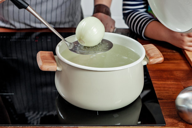 女人的手拿着一个带生洋葱的钢锅，把它放入沸水中。黑炉子上开着白锅。煮一个汤。图片下载