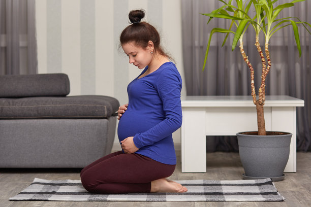 照片中，年轻的准妈妈正期待着孩子的出生，双手放在肚子上，跪在地毯上，深色的头发梳成发髻，与现代舒适的公寓内部形成对比。放松的概念图片下载