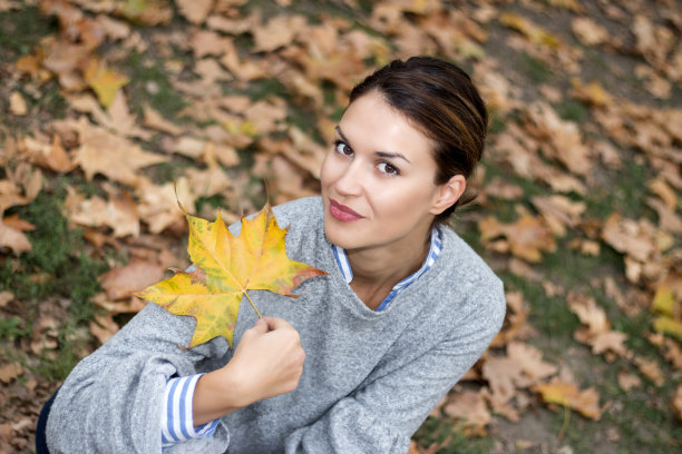 美丽的女孩坐在公园里的落叶上，微笑着，手里拿着一片黄叶。图片下载