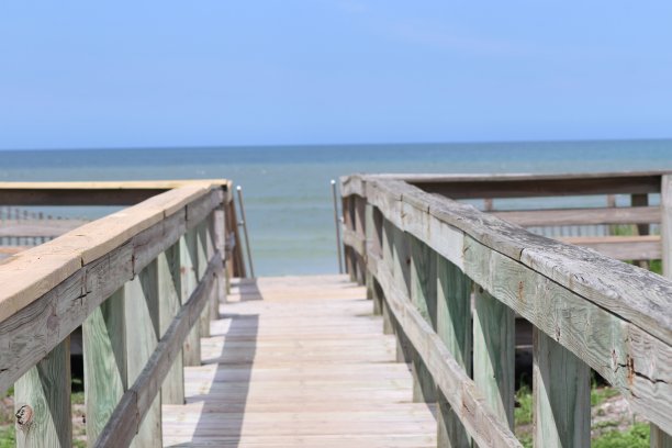 透视照片的木板路栏杆海滩进入地平线，蓝天，蓝绿色的海洋和绿色的草。图片下载