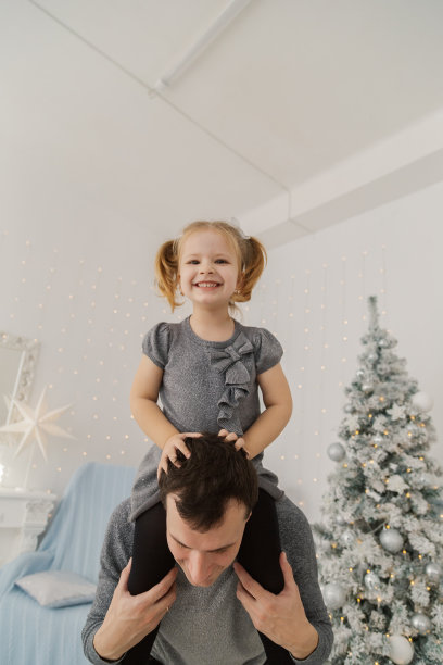 一个非常漂亮的女孩坐在她爸爸的肩膀上玩，在新年的房间里做鬼脸图片下载