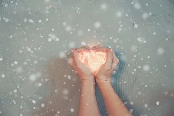 女人的手做成一颗心的形状，抱着温暖的圣诞彩灯，木制背景，俯视图图片下载