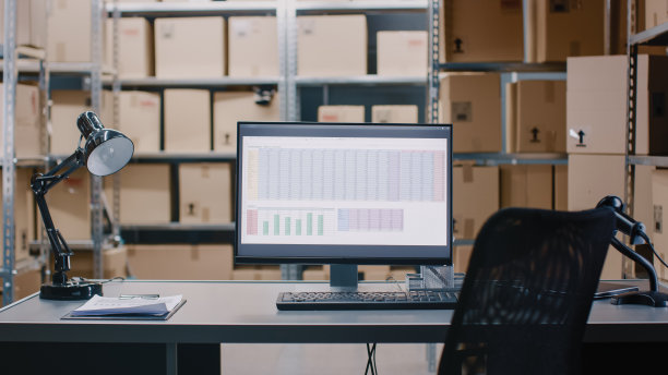仓库库存经理的办公桌和个人电脑的照片。在背景货架上装满了纸箱包装准备运输。图片下载