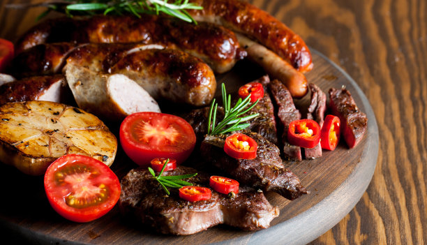 近距离的照片混合烤肉拼盘。牛肉，猪肉，家禽，香肠，烤大蒜，辣椒，红色的西红柿在木制的乡村背景。图片下载
