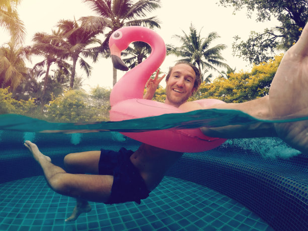 在度假村游泳池里玩充气火烈鸟的时候，一个快乐的男人在水下自拍。热带趣味度假概念图片下载