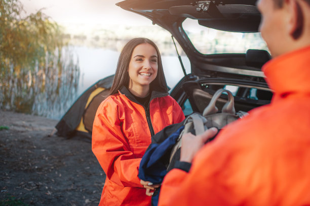 美丽开朗的年轻女子看着男人微笑着。她从他那里得到了背包。他们站在打开的汽车后备箱前。也有帐篷和湖的图片。图片下载