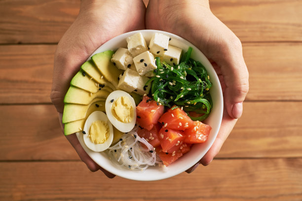 女士双手捧着盛有三文鱼、牛油果饭、丘卡沙拉、奶酪、鹌鹑蛋、白芝麻和黑芝麻酱油的碗，孤立在木质背景上俯视图片下载