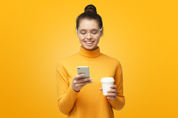 室内照片年轻漂亮的欧洲女人孤立在灰色背景拿着手机在手里，看屏幕微笑，浏览或阅读信息图片下载