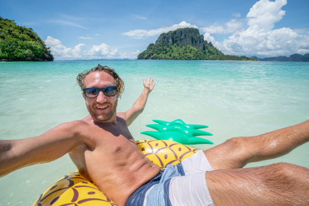 在泰国群岛清澈的海水中，一名年轻人在田园诗般的海滩上与充气菠萝自拍。人们旅行奢侈，乐趣和酷的态度概念图片下载