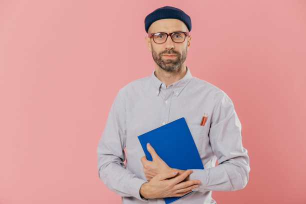 照片中，一名未刮胡子的年轻欧洲男子在镜头前看起来很自信，他有黑色的鬃毛，拿着记事本做记录，穿着正装衬衫，戴着帽子，粉色背景衬托着模特。图片下载