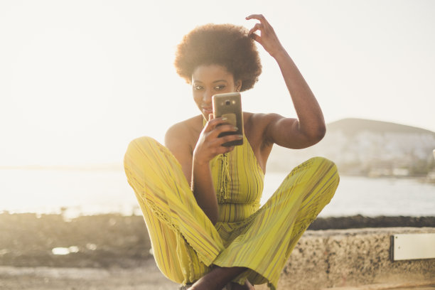 美丽的年轻黑人种族的非洲女孩有照顾和检查他的传统的非洲发型使用智能手机像镜子看到自己。千禧年现代人使用科技的概念图片下载