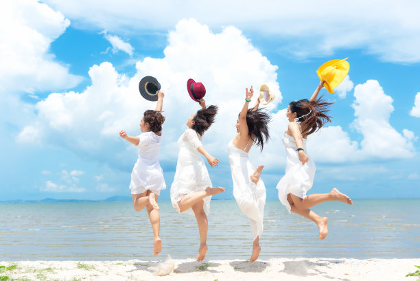 一群微笑的亚洲女子穿着时尚的白色连衣裙走在夏天的沙滩上，蓝天阳光的背景。快乐的女游客享受和放松图片下载