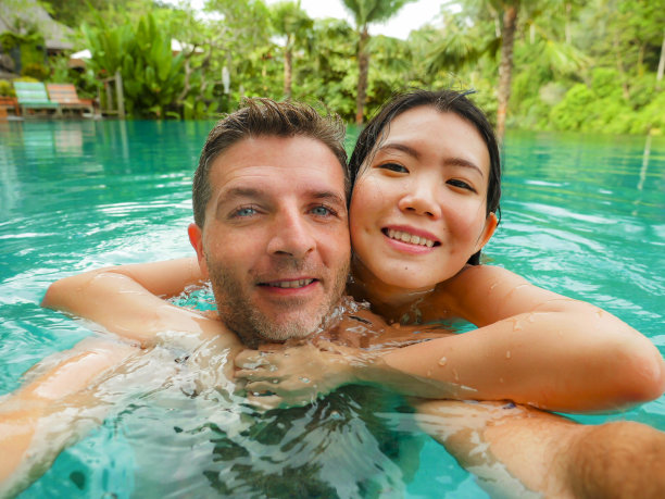 年轻快乐和美丽的混血亚洲女人和白人男人在热带度假胜地游泳池享受蜜月旅行庆祝爱情的浪漫自拍照片图片下载
