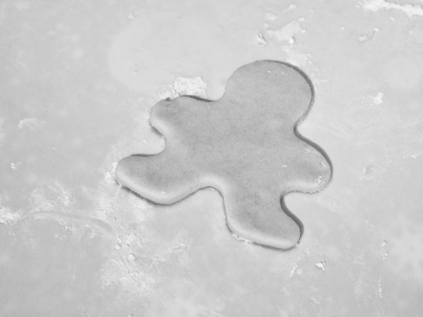 这是一张黑白的特写照片，上面是一块姜饼人形状的圣诞甜饼，用生的饼干面团切成，撒上面粉放在砧板上。图片下载