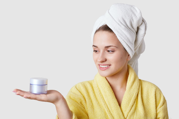照片横放着一个漂亮的年轻女子，面带温柔的微笑，有着健康的皮肤，手持塑料容器中的化妆品，穿着浴袍，模特们在白色的背景上，准备做面膜图片下载