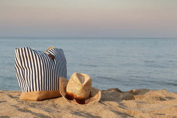条纹夏日包配草帽和太阳镜在沙滩上，以平静的大海为背景。日落时间。图片下载