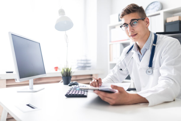 一个穿着白色长袍的年轻人坐在办公室的一张桌子旁。他手里拿着一支笔，目光直视。医生的肖像。图片下载