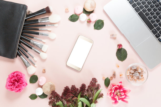 俯视图的办公桌，笔记本电脑，手机，绿叶，鲜花和杏仁饼干。平的。家庭办公室或美容博客工作空间。女性时尚配饰孤立在粉色背景上。图片下载