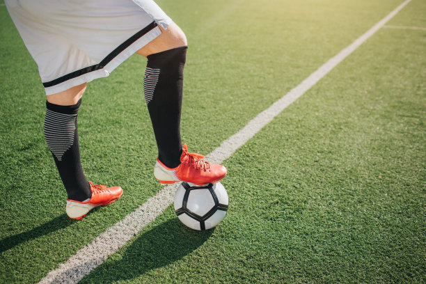 足球运动员下半身的图片。他站在绿地上，一条腿保持在球上。白线穿过绿色草坪。图片下载