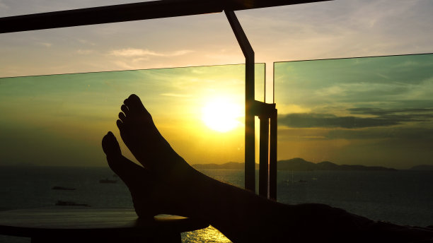 一个男人的脚的剪影放在一张木桌上，与美丽的下午日落在海边。图片下载