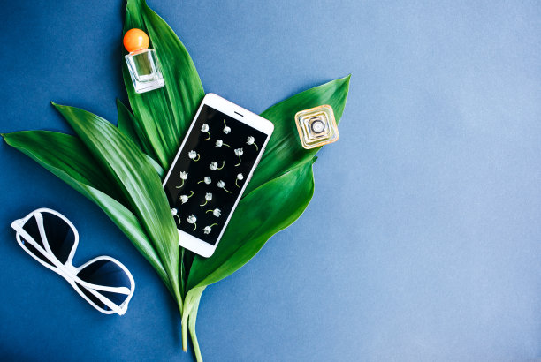 白色智能手机，花和绿色的叶子在蓝色的背景。山谷的百合极简平躺。母亲节或妇女节的礼物。本空间的地方。香水和太阳镜夏天旅行。图片下载