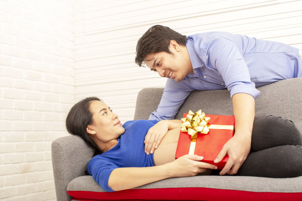 怀孕的母亲。给家人的礼物。微笑着的孕妇坐在沙发上，为她未出生的孩子准备礼物。父亲和母亲。妈妈和爸爸。亚洲人。图片下载