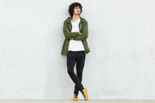 照片里的卷发男人穿着时尚的衣服，双臂交叉，目光远离，思考着什么，在工作室白色的墙壁上与世隔绝。人、风格、青春、自信理念图片下载