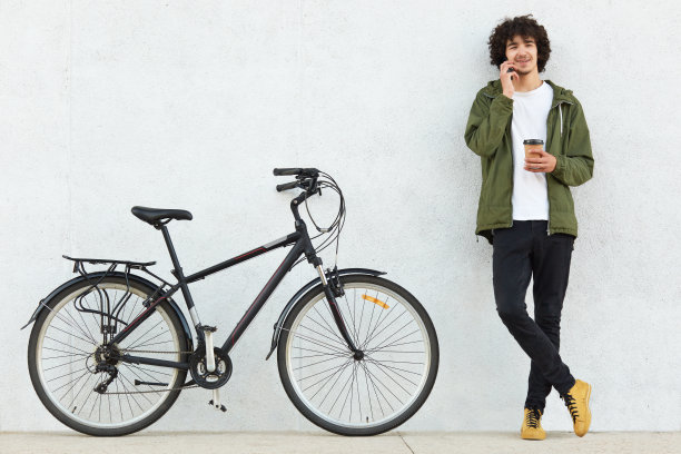 一头卷发的帅哥，穿着绿色的夹克，黑色的裤子，一头卷发，通过手机和女朋友聊天，拿着外卖咖啡，在自行车旁的白色背景下摆姿势图片下载